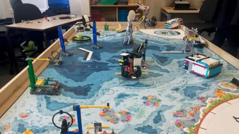 A large board with a sheet of paper designed to represent the ocean floor with a number of Lego items and models