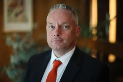 A man with short grey hair and wearing a suit with a red tie