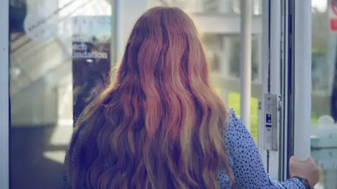 Una mujer camina por una puerta de un edificio de vidrio. Puedes ver la parte posterior de su cabeza, ella lleva un top azul con puntos negros y tiene un largo cabello rubio ondulado/rojo