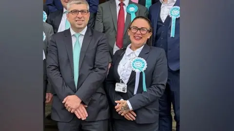 Kathryn Shaw stands next to council leader, Mark Arnull. They both wear suits and Shaw has a pale blue rosette indicating Reform UK on her jacket lapel. Other members of the Reform Party are stood behind them but you cannot see their faces.