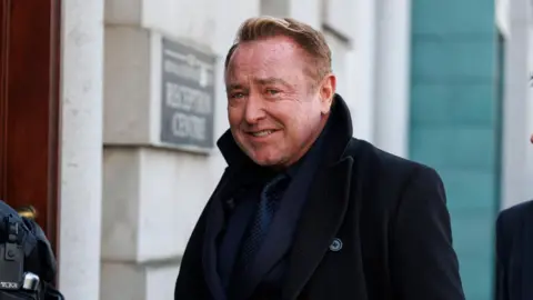 PA Media Michael Flatley, with short brown hair, wearing a navy suit, navy shirt, navy tie and black jacket. He is standing in front of a grey brick building. 