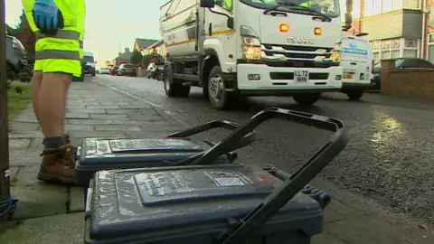 An image of a street in Newcastle where food waste caddies have been placed on the kerbside ready for collection. A bin lorry is approaching a binman who is waiting to tip the caddy contents into the back of the truck