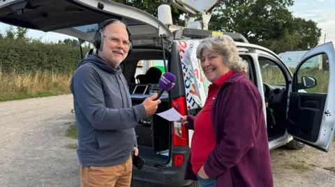 Danny Pike wears a navy blue hooded jumper and black headphones speaks to farmer Sarah who wears a red polo shirt and purple fleece in front of the sign-written BBC Sussex radio car.