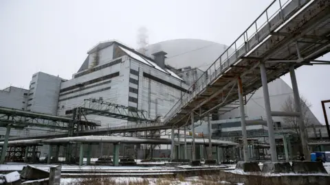 EPA A gray industrial building with various pipes, walkways and platforms attached to it stands in front of a large dome-shaped building of the same color. The ground is covered in snow and the steel buildings show signs of rust and age.