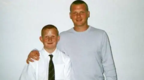 White family A man in his 20s with his hand on the right shoulder of a schoolboy wearing a white shirt and striped tie. The man is wearing a grey sweatshirt. Both are smiling. The image has been taken against a white wall.
