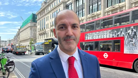Man wearing a Royal blue suit, white shirt and red tie, on a high street with a red London bus passing by it, along with other traffic.