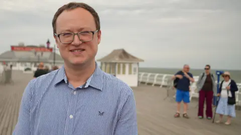 Shaun Whitmore/BBC A man in a white and blue striped suit smiles at the camera. He has short brown hair and is wearing glasses