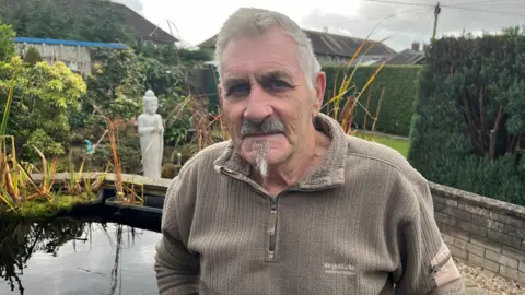 Koi enthusiast Steven Smith stands in front of his Carp pond. The water is behind him, with reeds growing high, a stone wall built around it and a Buddha statue in the background. He is wearing a light brown sweater