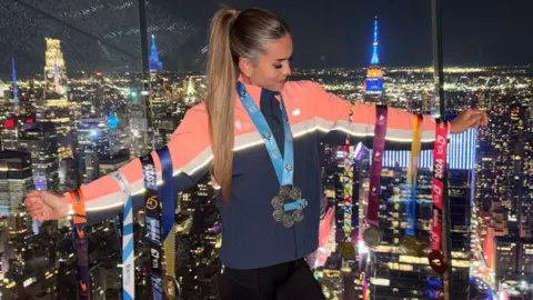 Lucy Scott Lucy Scott olha para baixo enquanto pendura as medalhas da maratona nos braços. Ela tem um longo rabo de cavalo marrom e usa um paletó azul e rosa. As cores variam entre ouro, prata e bronze e apresentam fitas de diversas cores. Ela está em frente a uma janela que mostra o horizonte da cidade de Nova York à noite. 