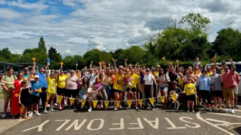 Naomi Weir A group of women, men and children cheering, many with their arms aloft and some are holding yellow bunting. Behind them are trees and blue and white skies. 