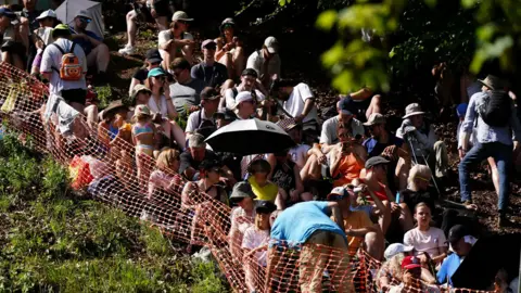 PA Media Spectators seeking shade beneath the trees at the Gloucester cheese rolling event. There is a large crowd of people sitting down on the grass behind an orange plastic barrier, wearing sunglasses, hats and carrying umbrellas.