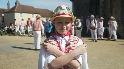 Jamie Niblock/BBC Tyler is wearing his Morris dance costume. He is wearing a beige-coloured straw hat with various badges on it. He is holding two white handkerchiefs, crossing his arms and smiling. In the background, across a square, are other dancers and houses.