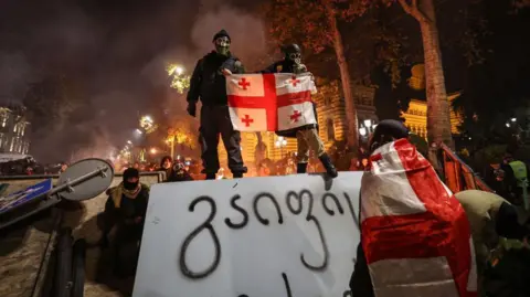 GIORGI ARJEVANIDZE/AFP Seorang pengunjuk rasa yang mengenakan bendera Georgia berdiri di barikade saat bentrokan dengan polisi pada 1 Desember 2024