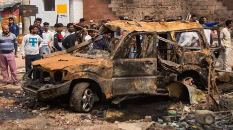 Os espectadores da Getty Images se reúnem em torno de um veículo carbonizado após os distúrbios de 2020 Delhi 