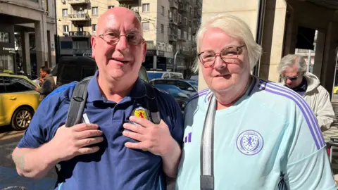 Gavin Keith and Edith Stewart looking straight at the camera. They are both wearing scotland shirts. Gavin is on the left. He is bald and is wearing glasses. Edith is on the right and has white blonde hair. She is also wearing glasses.