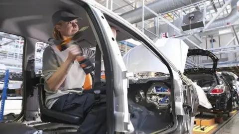 A woman wearing a grey shirt, drilling into the chassis of a car