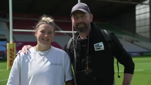 Ella Cromack, wearing a white T-shirt, is standing in front of rugby posts. Her dad Simon is wearing a cap with Harlequins on it and is wearing a black hooded top with "Give Simon Time" written on it. He has a dark beard and has sunglasses tied around his neck. 
