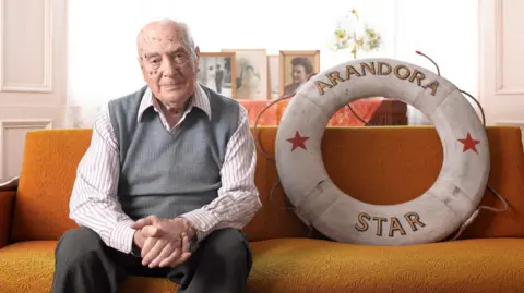 An elderly man in a striped shirt, grey tank top and grey trousers sits on a sofa next to a life buoy ring with the name Arandora Star on it