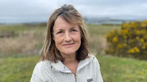 A woman with shoulder length blonde hair is smiling into the camera. She is wearing a cream fleece zip jacket. The background is blurred, but she is standing outside in a grassy area with yellow flowers to one side. 