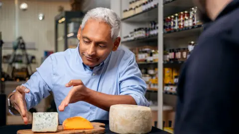 Scott Barron A man with grey hair in a blue shirt points at a block of cheese