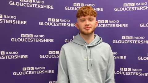 A a ginger-haired young man in a grey hoodie standing in front of a purple BBC Radio Gloucestershire branded backdrop with white writing on it