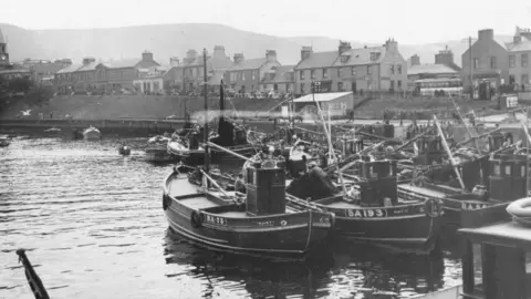 Getty Images A black and white picture of Girvan, with rows of fishing boats docked by the pier