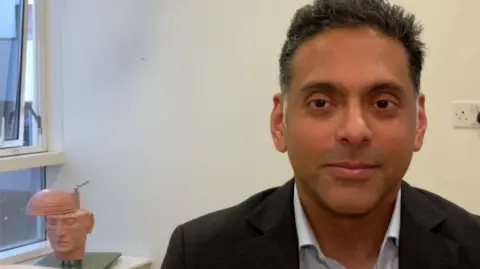 Ryan Mathew, who is associate professor at the University of Leeds and honorary consultant enurosurgeon at Leeds Teaching Hospitals NHS Trust. He has dark brown and grey hair and is wearing a suit. 