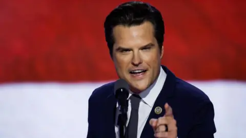 A close up of Matt Gaetz on stage at the Republican convention in Milwaukee, Wisconsin in July. He is speaking into a microphone and is pointing with his hand. He has dark brown hair and wears a dark suit, white shirt and navy tie.