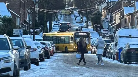 Casey Buckley A bus at a sideways angle with snow on the ground. Vehicles are parked either side of the road