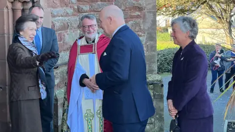 Princess Anne, who is wearing brown skirt, jacket and leather gloves, holds her hand out to Lieutenant Governor Sir John Lorimer, with the Dean of the Cathedral Nigel Godfrey, Chief Minister Alfred Cannan and Lady Lorimer also standing by.