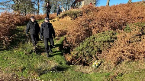 Two police officers, wearing black clothing and caps, walk along a grassy slope. A number of other people are behind them on higher ground. A stone building is also visible.