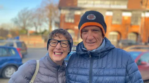 A woman wearing a grey puffer coat. She has short, curly hair poking out from under a beanie hat. She's wearing glasses. She's standing next to a man in a blue puffer coat. He is wearing a blue and orange Luton Town football club beanie. They are standing in front of a road.