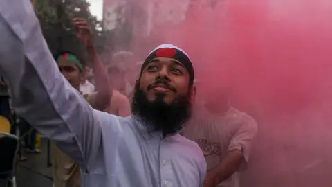 NurPhoto via Getty Images A bearded young man in a blue shirt stands waving a can making red smoke in amongst a crowd, wearing a green headband with a red dot in the middle - a version of the national flag. Blurry people in a crowd can be seen in the background