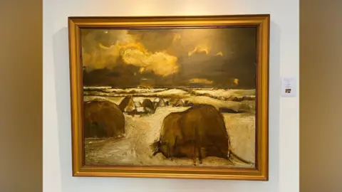 Haystacks in Winter hangs on a white wall in a golden frame. It is a snowy scene with huts dotted around the flat landscape under a heavy sky.