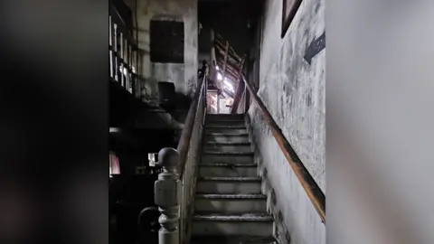 Images shows inside a pub after a fire. The open staircase, walls and both the ground and upstairs show smoke damage with daylight coming in part of the roof damaged by the blaze.