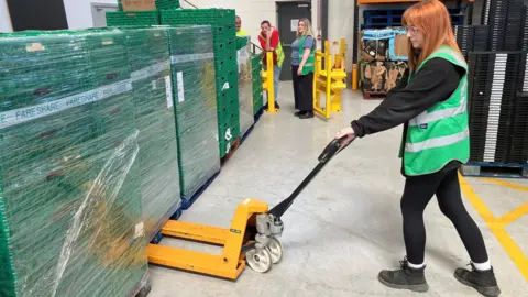 A FareShare worker moves palettes at the Middlesbrough hub. She is wearing black leggings and long-sleeved top, as well as a green vest. Her hair is straight and ginger. 