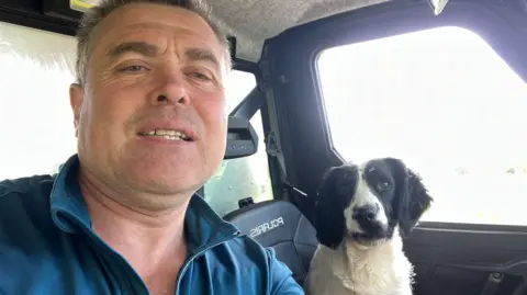 Thomas Conway A man is pictured in the front seat of a jeep, with a springer spaniel dog sitting in the passenger side seat. the man is wearing a blue jumper. the dog is black and white in colour.