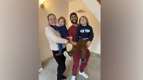 Billie Van Der Walt A woman holds a child, smiling at the camera. Next to her a man holds a toddler. Both are smiling as they stand in an empty living room of a house with grey carpet.