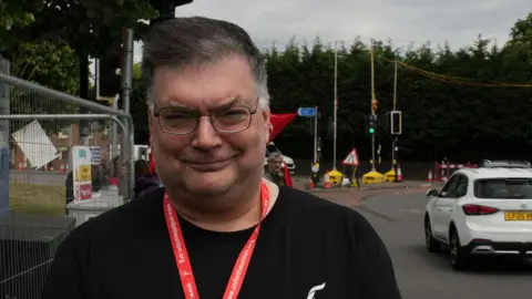 John Fairhall/BBC Paul Hardingham outside Addenbrooke's Hospital in Cambridge. Has greying black hair and is wearing glasses and a black T-shirt. Behind him can be seen roadworks and a roundabout.