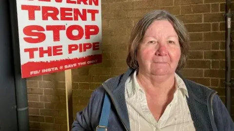 Ludlow mayor Beverley Waite wearing a light-coloured blouse and blue jacket in front of a placard reading: "Severn Trent stop the pipe"