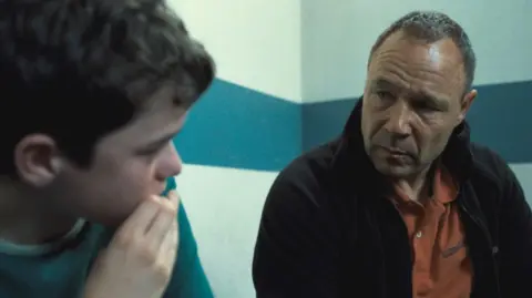 Still of Owen Cooper and Stephen Graham in Adolescence. Stephen Graham is wearing an orange uniform shirt under a black fleece. Owen Cooper is wearing a green shirt. They are both looking at each other.