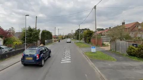 Google Google street view screenshot of a residential road with semi-detached houses and large gardens on both sides. Two cars and a motorcycle are driving on the road and it is a cloudy day.