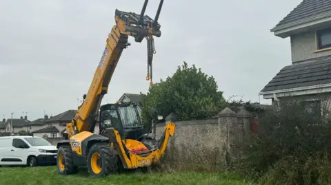 A JCB and a white van parked on a patch of grass. There is a grey wall behind the JCB and homes in the background. 