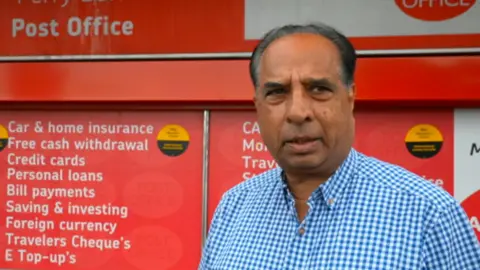 Roshan Dall, postmaster at Perry Barr Post Office, stands outside the front of the Post FFICE WITH RED AND WHITE POST OFFICE BRANDING BEHIND.