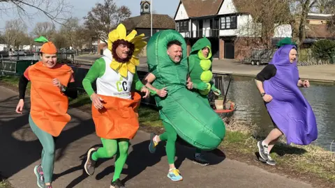 Tom Williams/BBC The group dressed as their respective vegetables and the sunflower running along a river. They have smiles on their faces as they run.