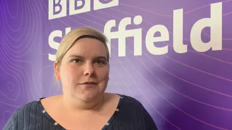 BBC/Mick Lunney A woman with blonde, tied-back hair stands in front of a BBC Sheffield logo board.