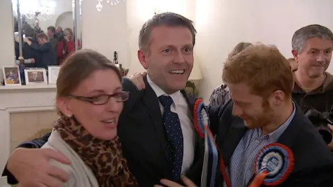 Philip Ozouf is celebrating being elected. He is being hugged by his partner, Kevin, to the right and a woman to his left. Ozouf is wearing a navy suit with a navy spotted tie and a blue, red and white rosette. He's in a living room with people behind him.