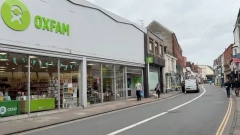 Robby West/BBC The street with Oxfam on the left side - it is a large shop with six big glass panels at the front and the green Oxfam sign. You can see shelves and tables with cards, books and other items on.