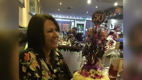 Maria Britton on her 50 birthday, in a restaurant with a 50 balloon and a birthday cake. She is smiling and wearing a floral top.