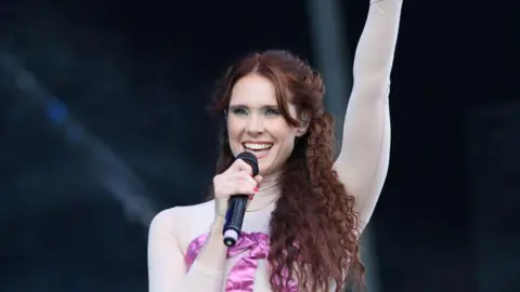 Getty Images Kate Nash who has long red hair singing with a microphone. She is wearing a pink dress